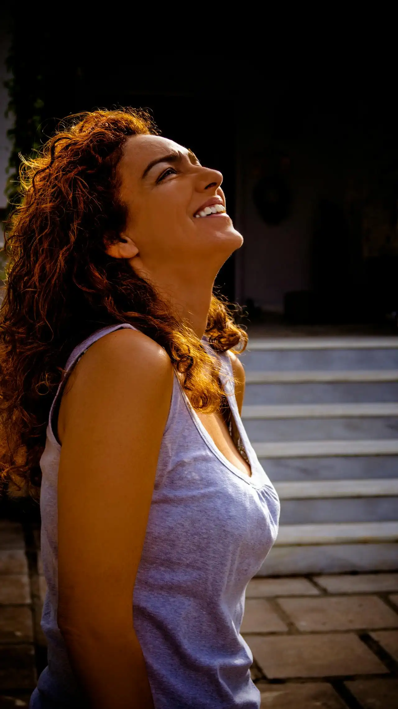 Mulher com cabelos cacheados ruivos sorrindo ao ar livre, em posição de perfil, usando camiseta regata clara. Luz do fim de tarde destaca o cabelo e o rosto.