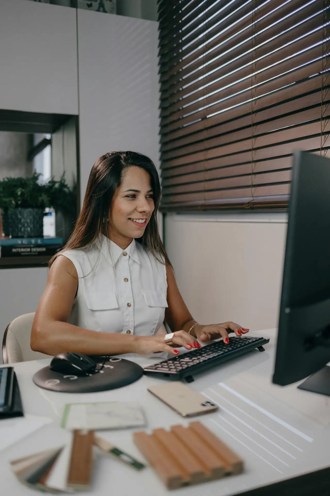 Mulher usando computador em ambiente de escritório, sorrindo enquanto digita no teclado; ao lado há mouse, documentos, amostras de cores e um cartão, com luz natural e persianas ao fundo.
