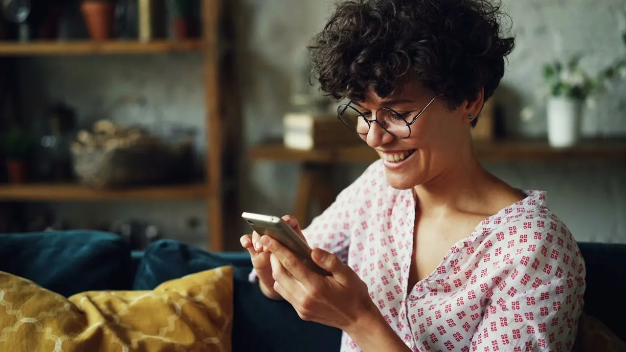 Mulher jovem com cabelo cacheado e óculos sorrindo enquanto usa o celular em casa, em ambiente aconchegante e desfocado ao fundo.