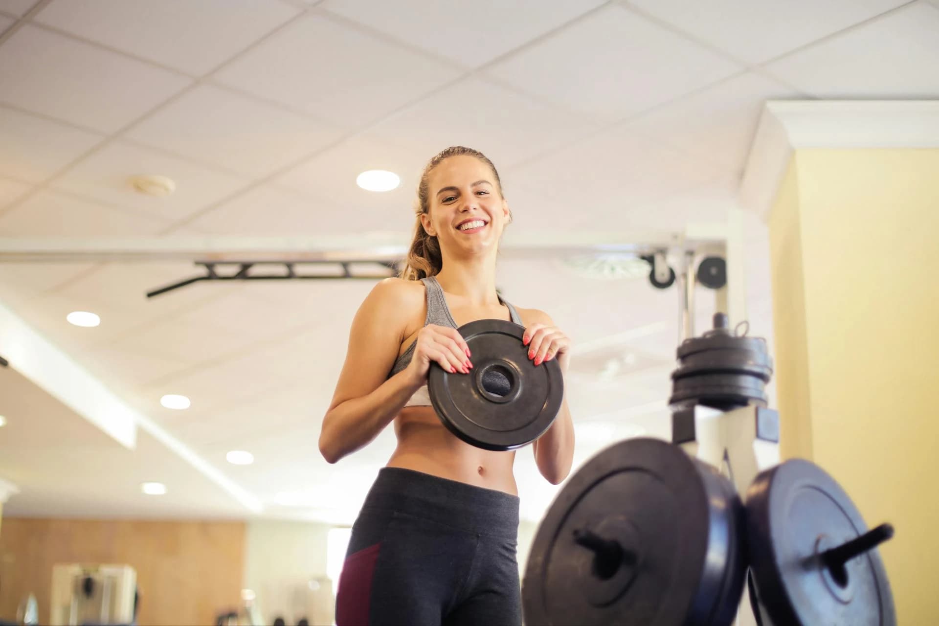 Mulher treinando na academia com anilha de musculação, segurando um peso em formato circular enquanto sorri. Ambiente de treino com outros discos ao fundo e iluminação do salão.